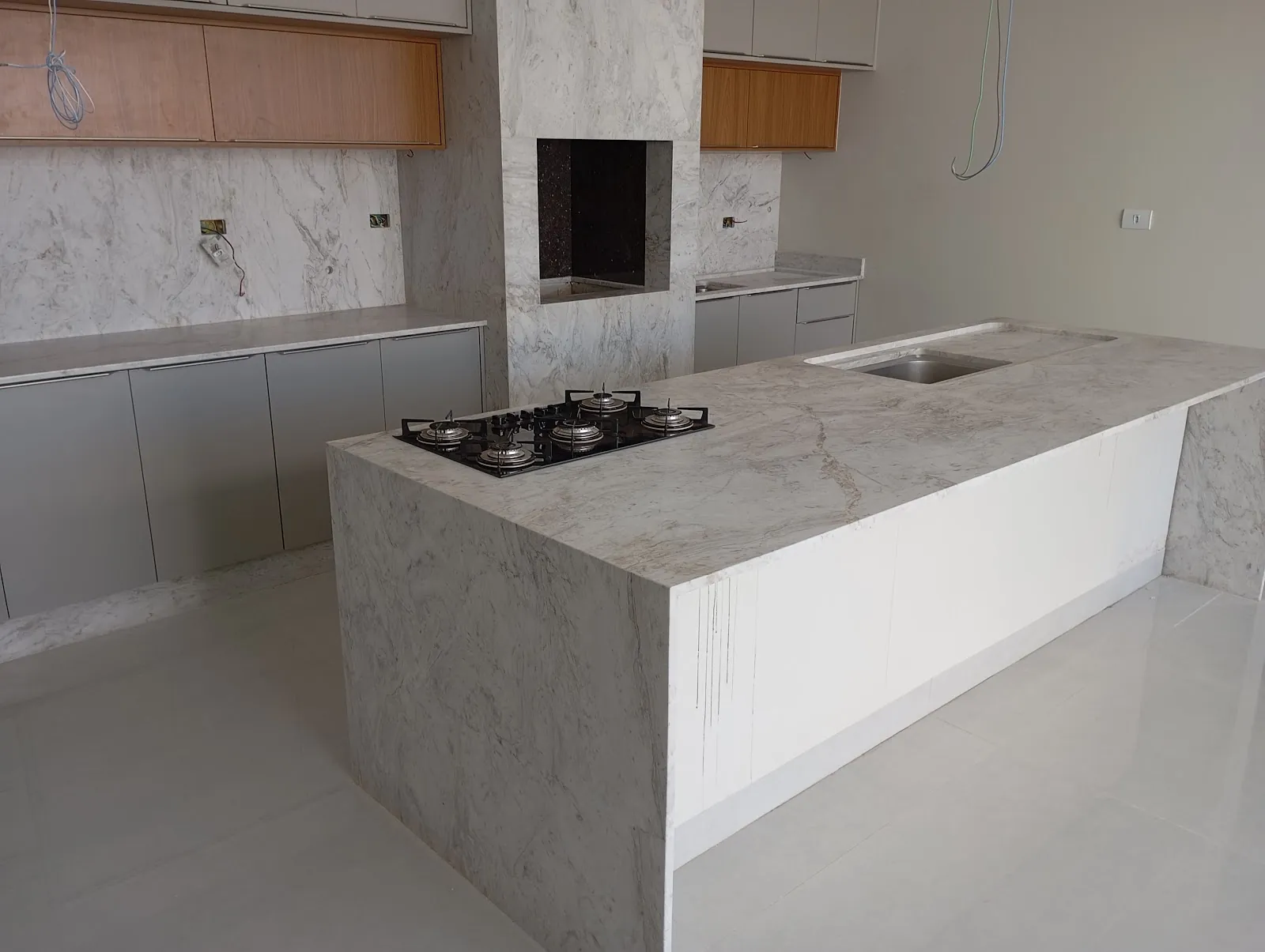 Ilha em mármore claro com cooktop e lateral em cascata, com churrasqueira ao fundo, feita pela Marmoraria Stone House em Maringá